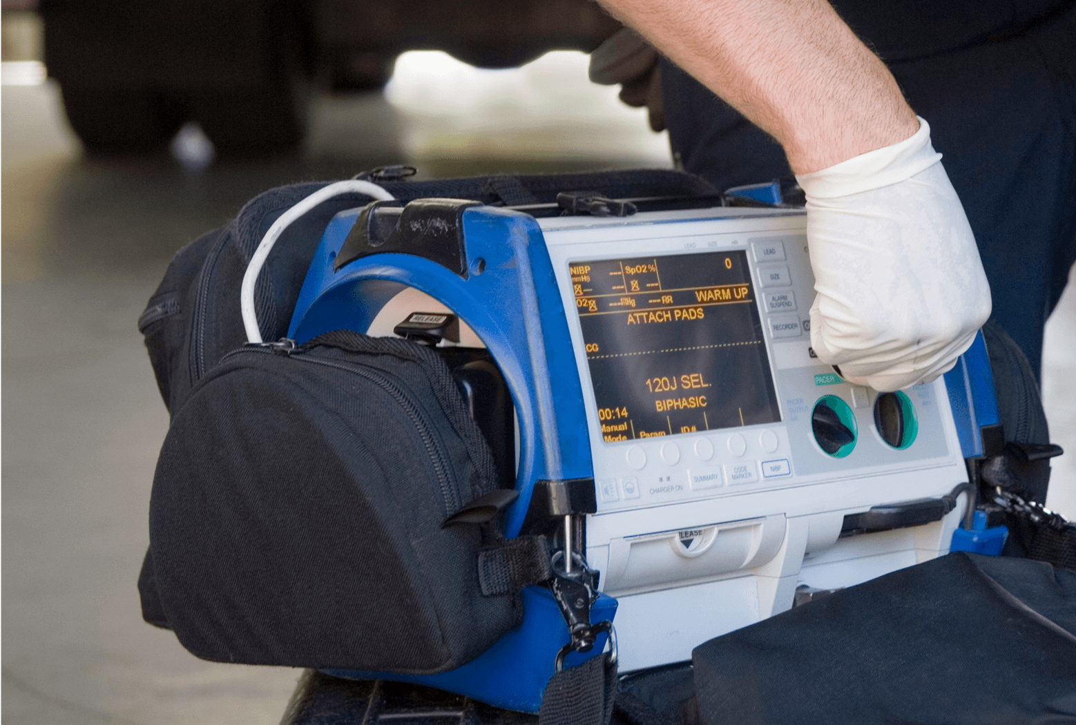 Close-up of a medical professional's gloved hand turning a green knob on a portable defibrillator and patient monitor.
