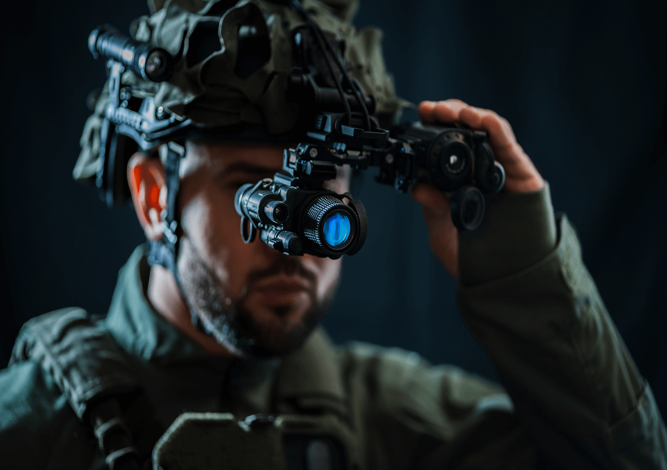 A soldier in tactical gear adjusts a knob on his helmet-mounted night vision goggles in a dark environment.